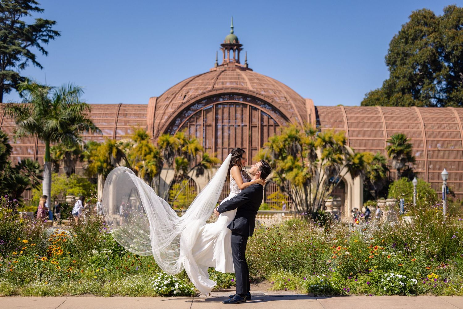Couple embraces in front of historic domed building while bride's veil flows dramatically in the breeze.