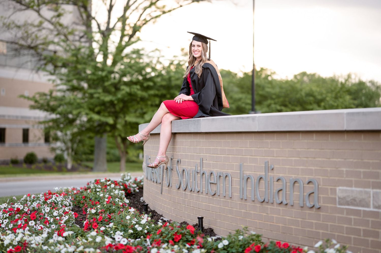 Graduate in cap and gown sitting on a brick wall with red flowers blooming below.