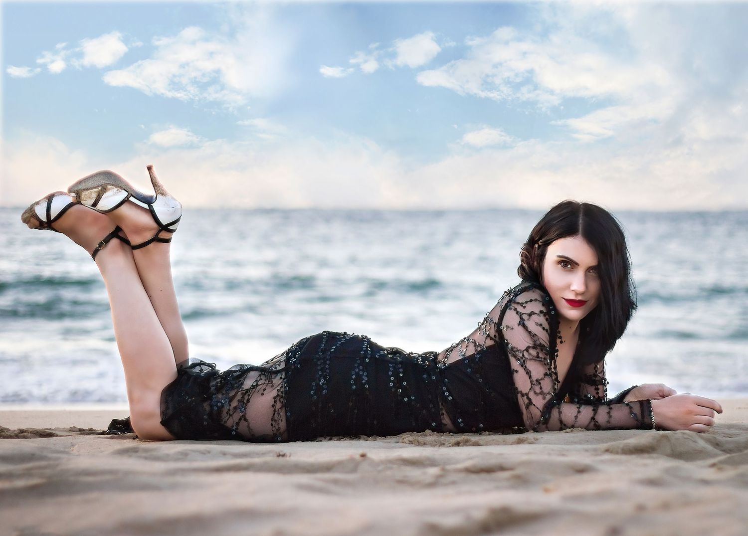 Dark haired woman in beaded evening gown, high heels, laying on her front on the beach with ocean in background.