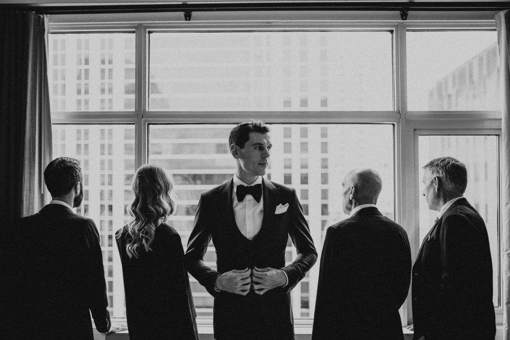 Silhouette of wedding party gathered by large window in formal attire.
