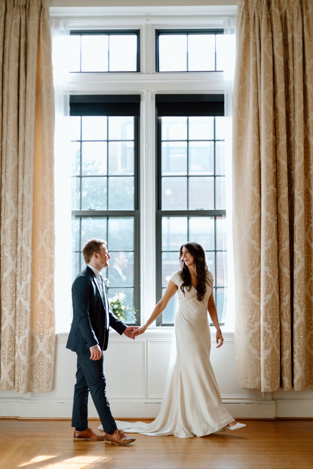 Bride and groom hold hands in front of tall window with elegant curtains.