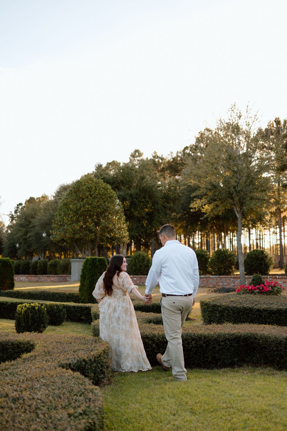 A couple walks hand in hand through a manicured garden with ornamental hedges at sunset.
