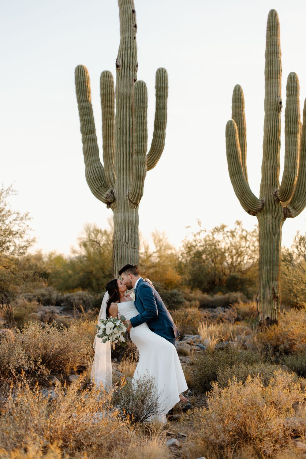 Romantic wedding couple embraces among towering saguaro cacti in desert landscape at sunset.