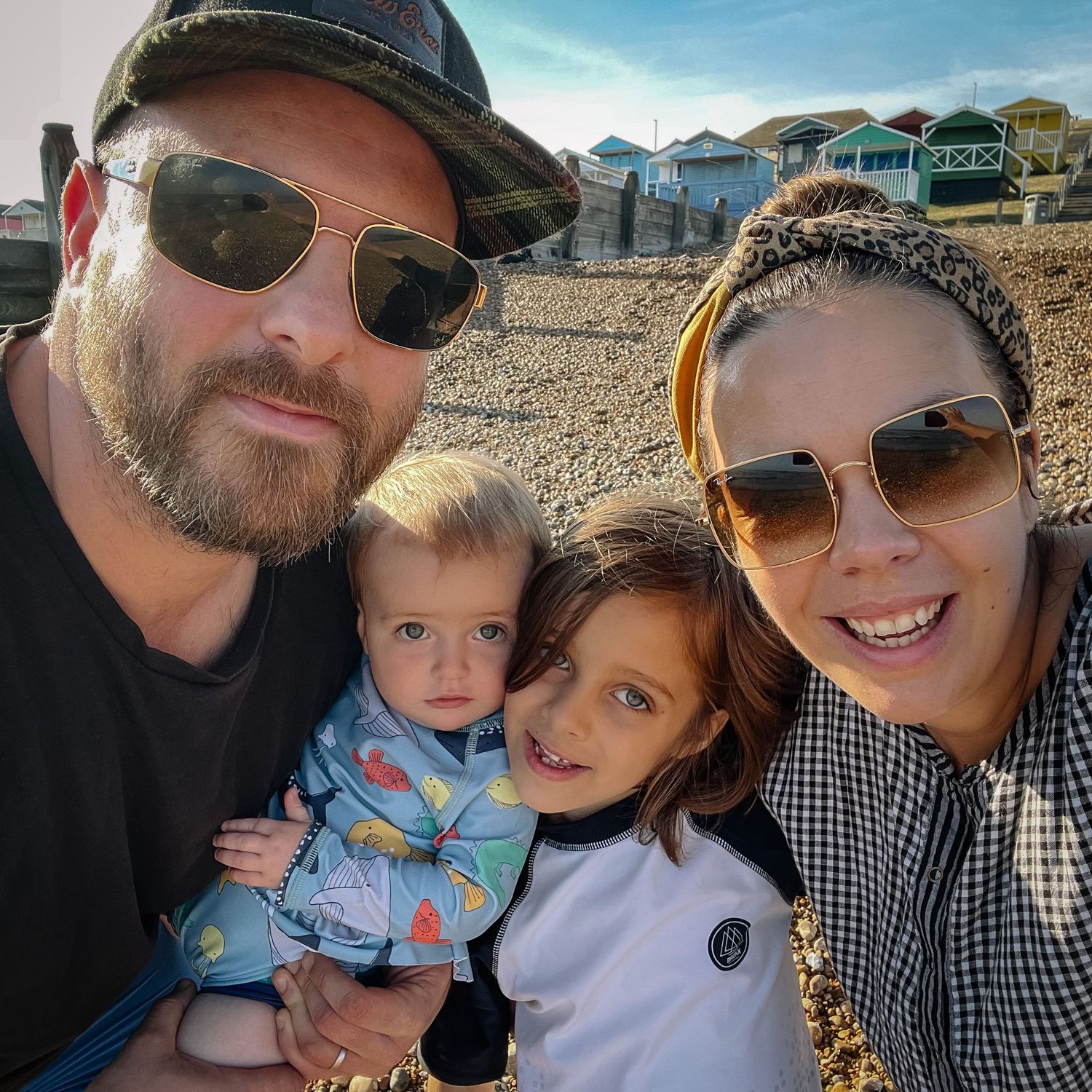 Family selfie at beach with houses in background on sunny day.