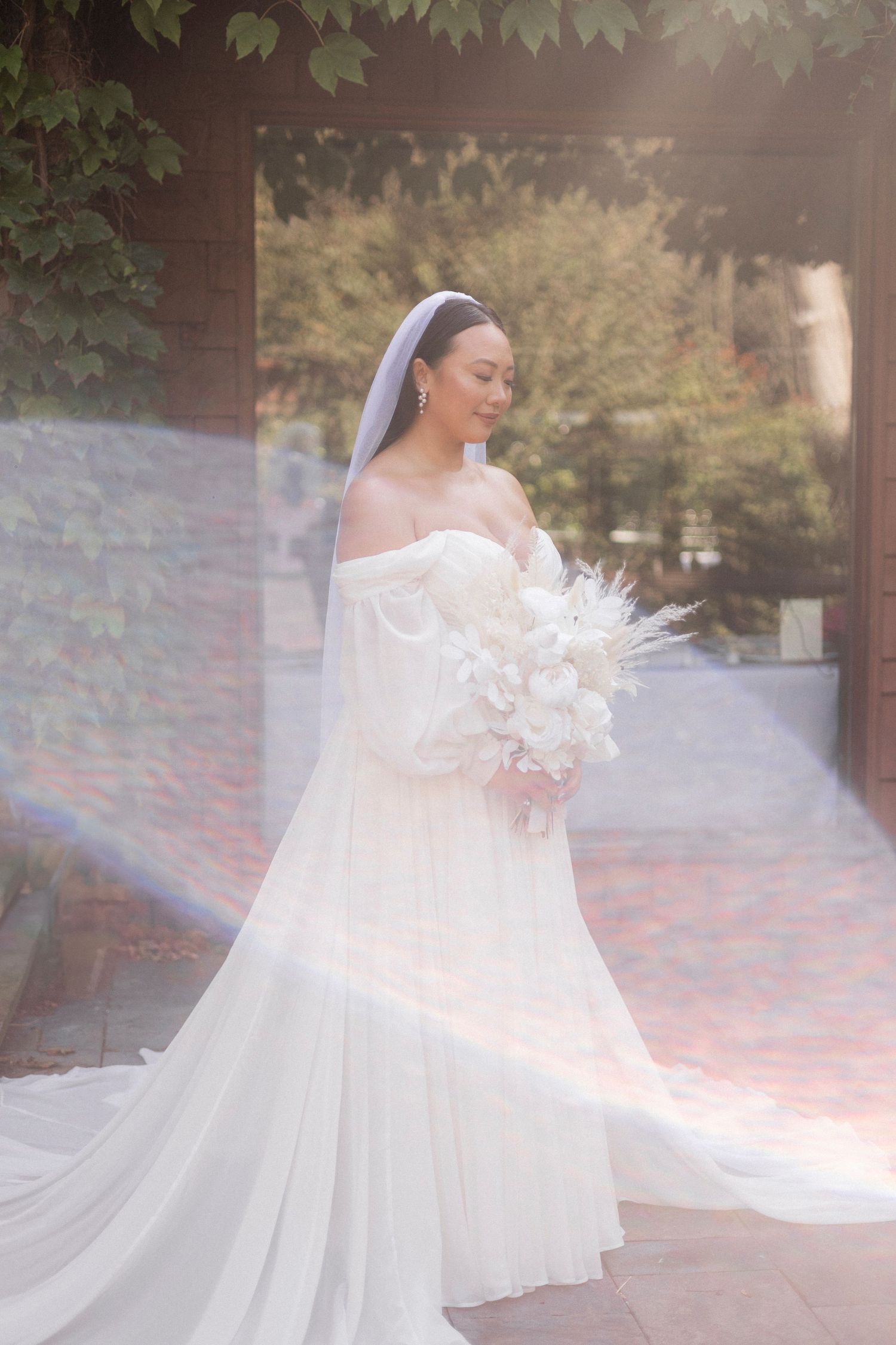 Elegant bride in an off-shoulder white gown holds a bouquet in a dreamy outdoor setting.