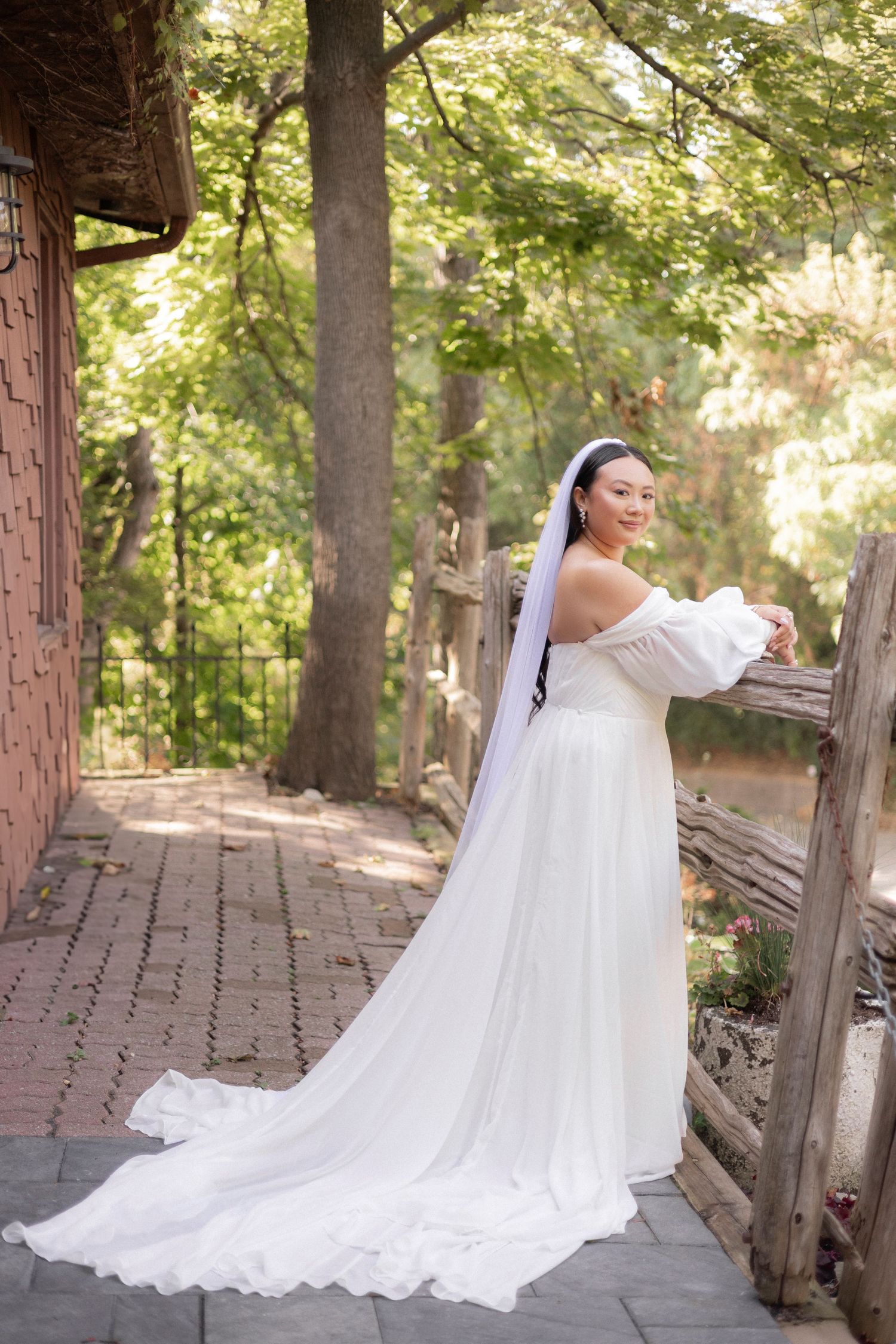 A bride in white gown poses on brick pathway under trees with trailing veil.