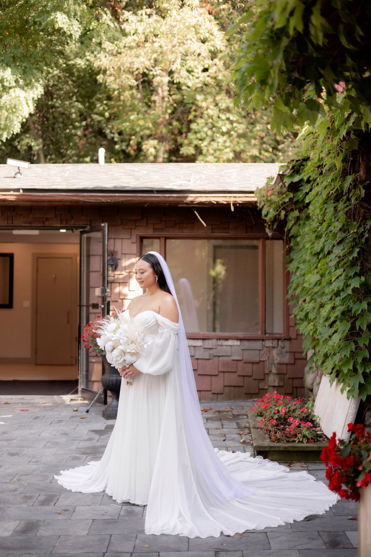 A bride in white gown stands beside a brick building surrounded by greenery and flowers.