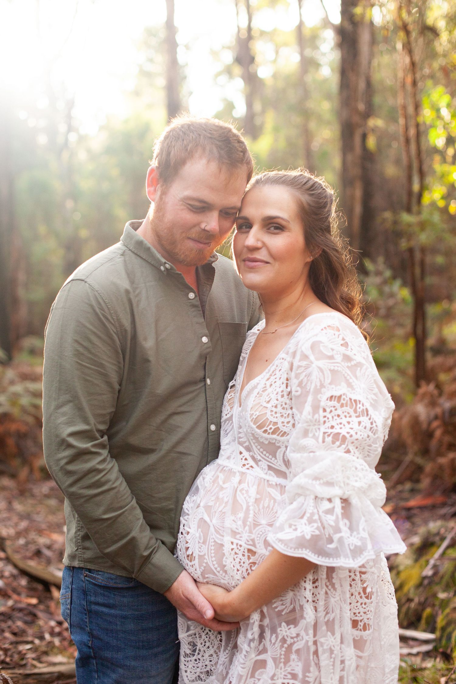 Expectant couple in woodland setting with sunlight, wearing white lace dress.