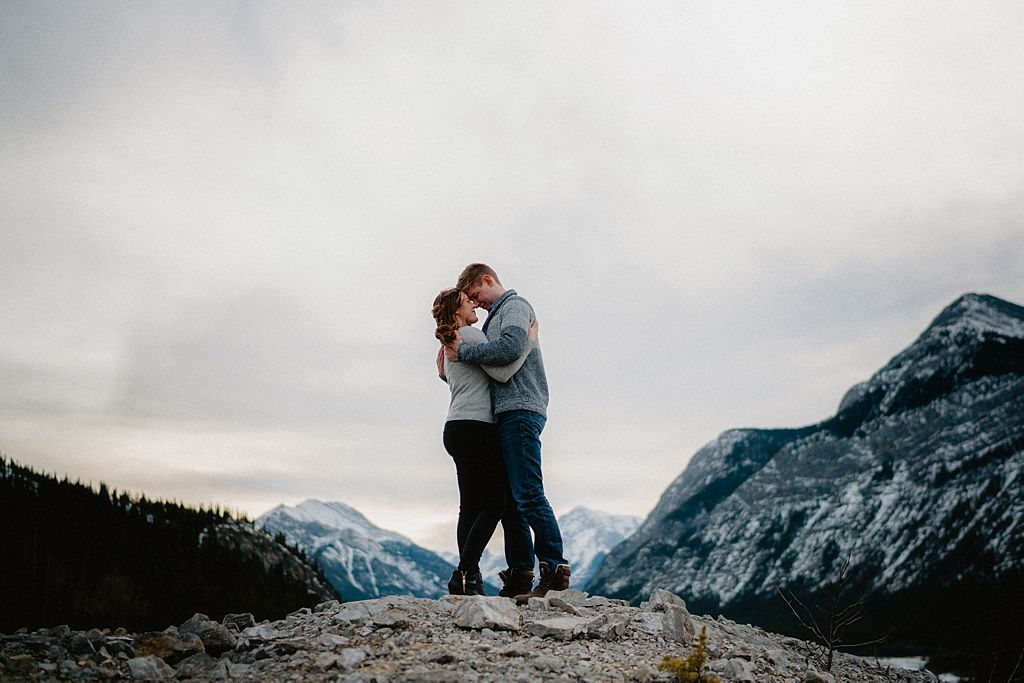 Couple standing close on rocky outcrop with snow-capped mountain peaks in background.