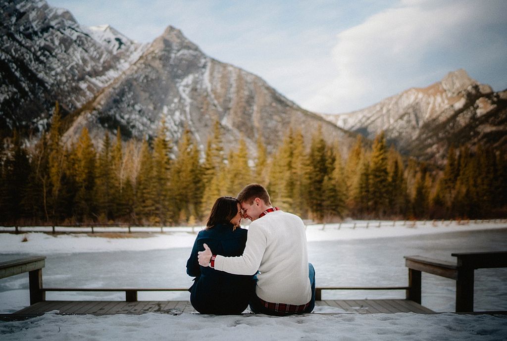 Couple sitting on wooden bench overlooking snow-covered mountain landscape.