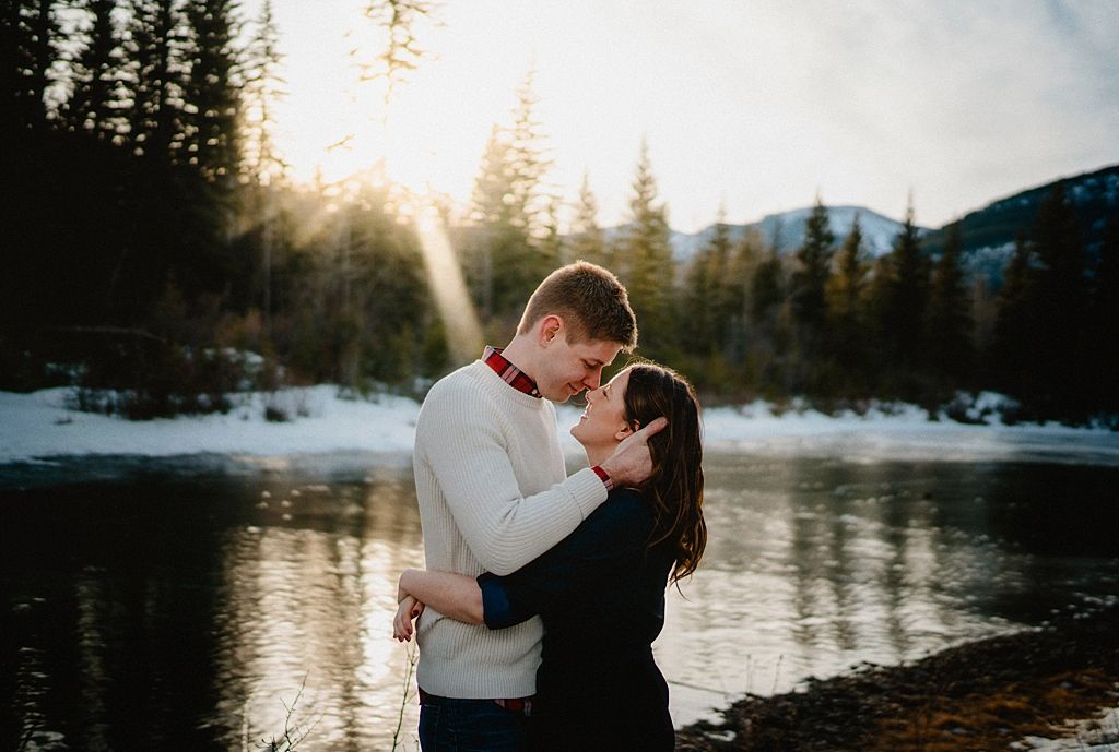 A romantic moment shared between a couple by a snowy mountain lake at sunset with evergreen trees in the background.