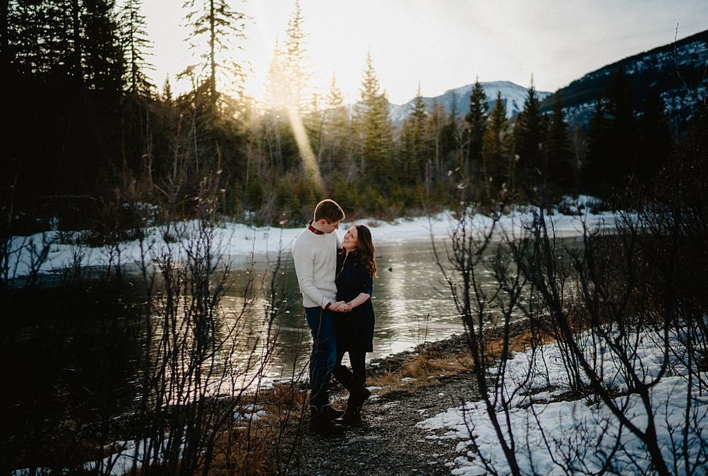 Silhouette of a couple embracing by a snow-covered lake with sunbeams filtering through mountain pine trees.