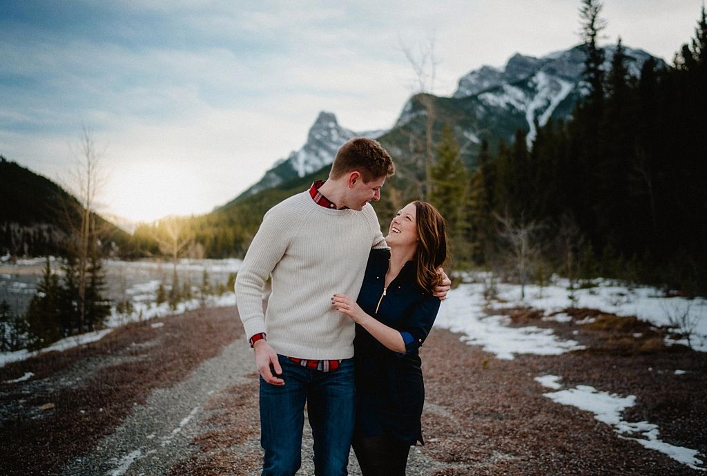 Winter engagement photos with couple standing on snowy path with mountain peaks in background.