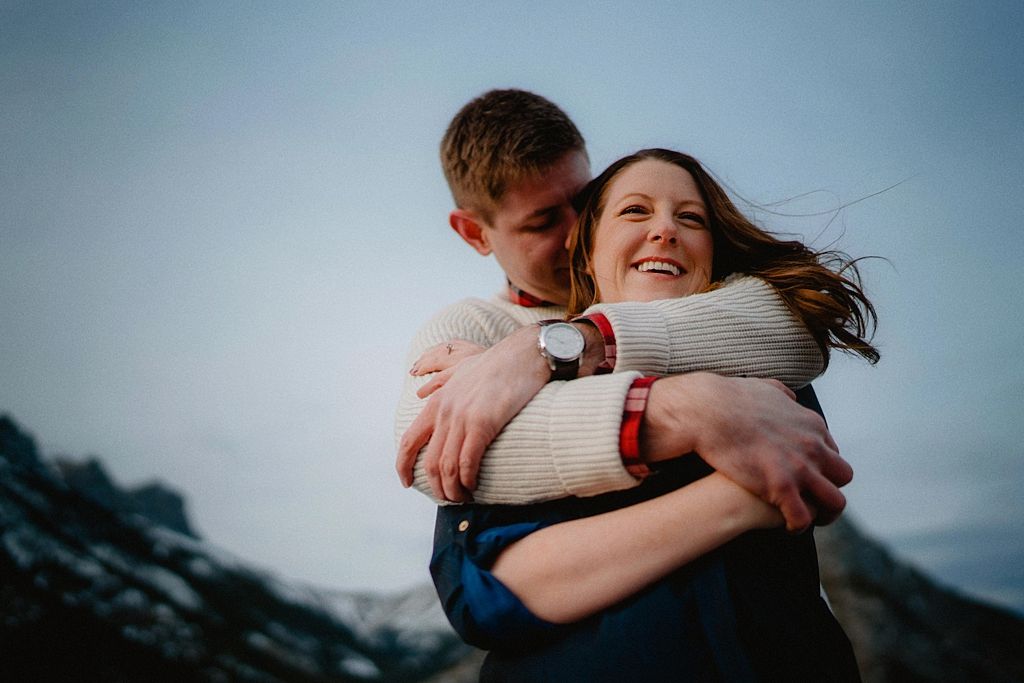 Candid embrace captured against snow-capped mountain backdrop during winter photoshoot.