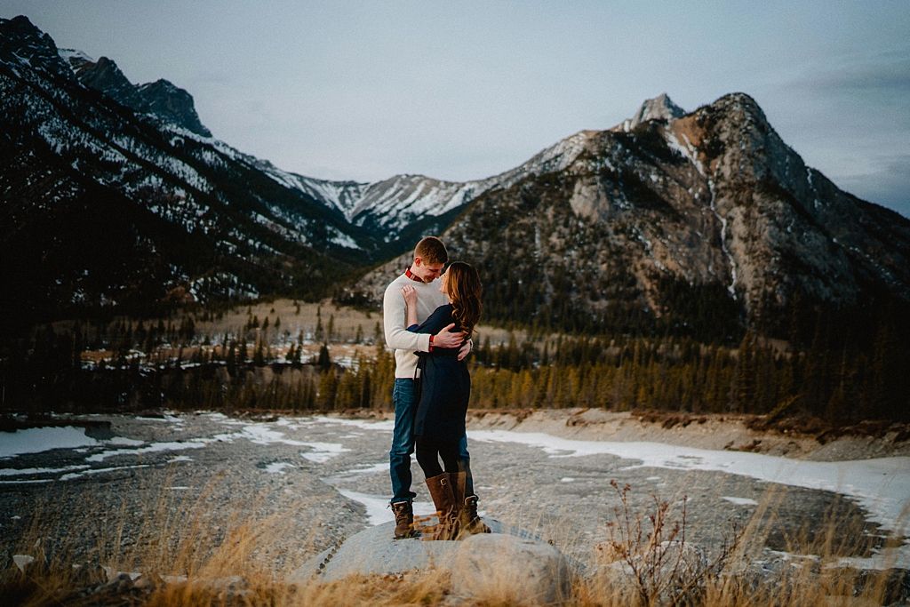 Winter engagement photo with couple embracing on mountain overlook with dramatic peaks behind.