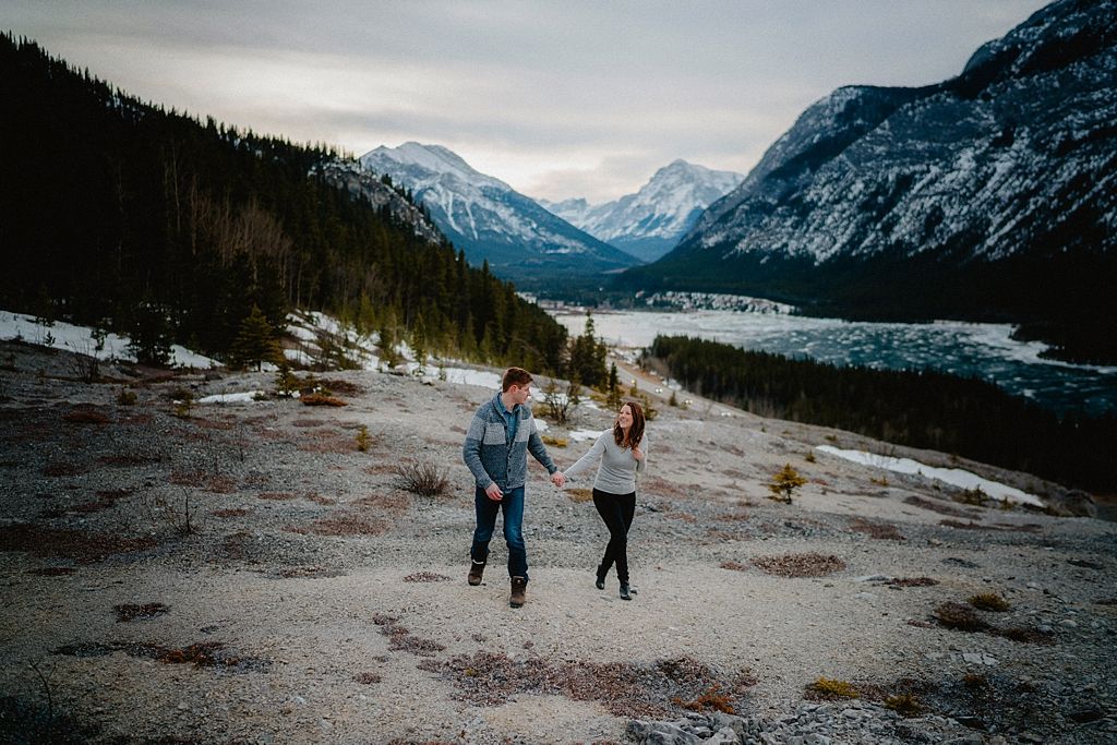 A couple runs joyfully along a rocky terrain with snow-capped mountains and evergreen forests in the background.