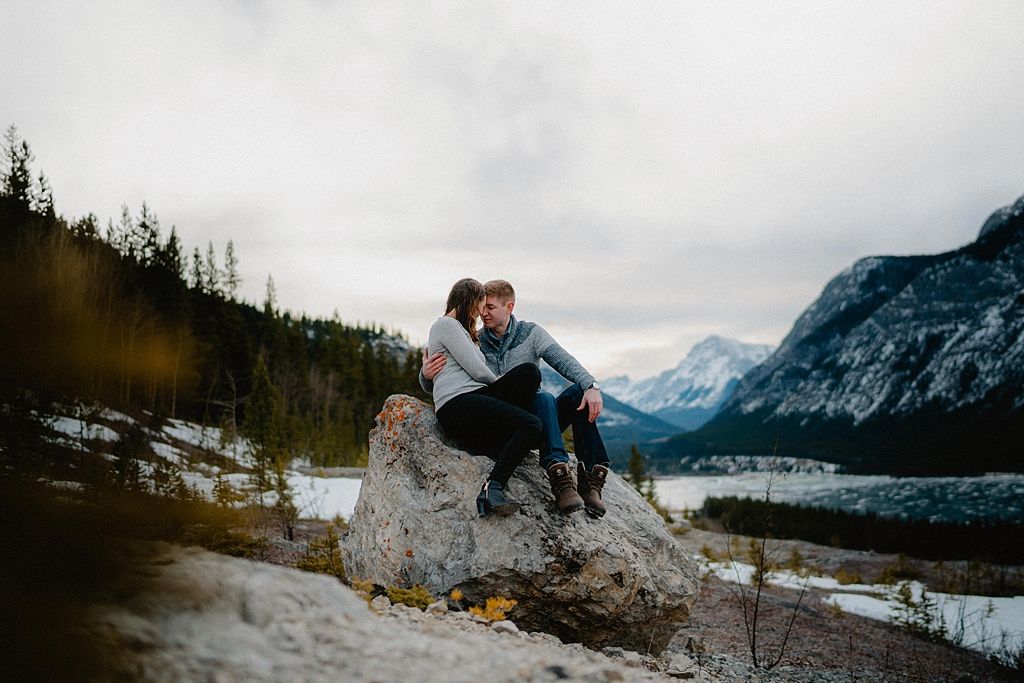 Two people share a moment while sitting on a large boulder overlooking a mountainous winter landscape.