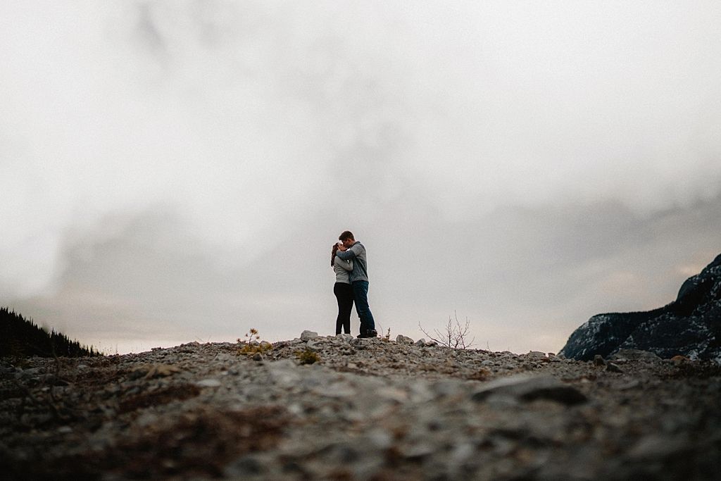 A silhouetted couple embraces on a mountain ridge against a moody sky.
