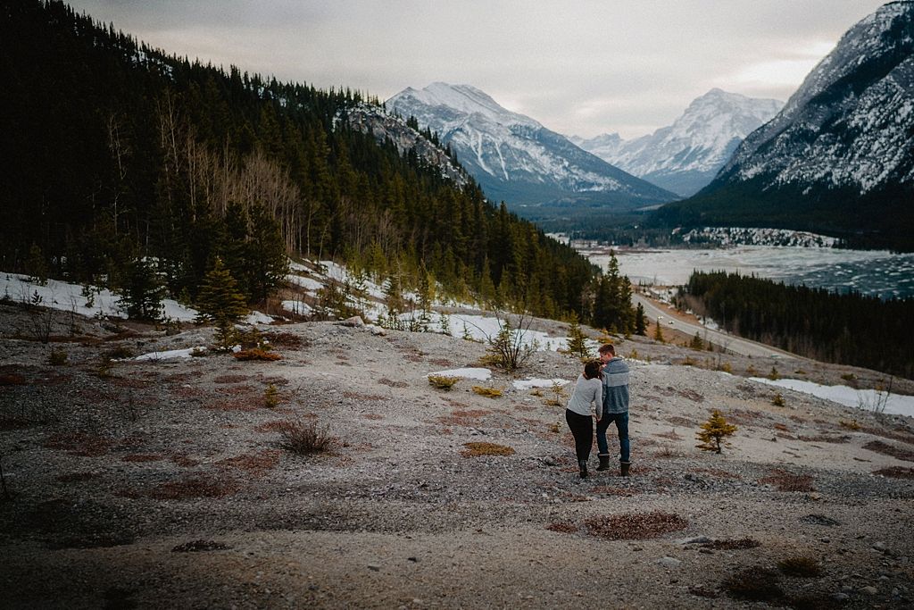 Two people stand together on rocky terrain with snow-dusted mountains and valley views.