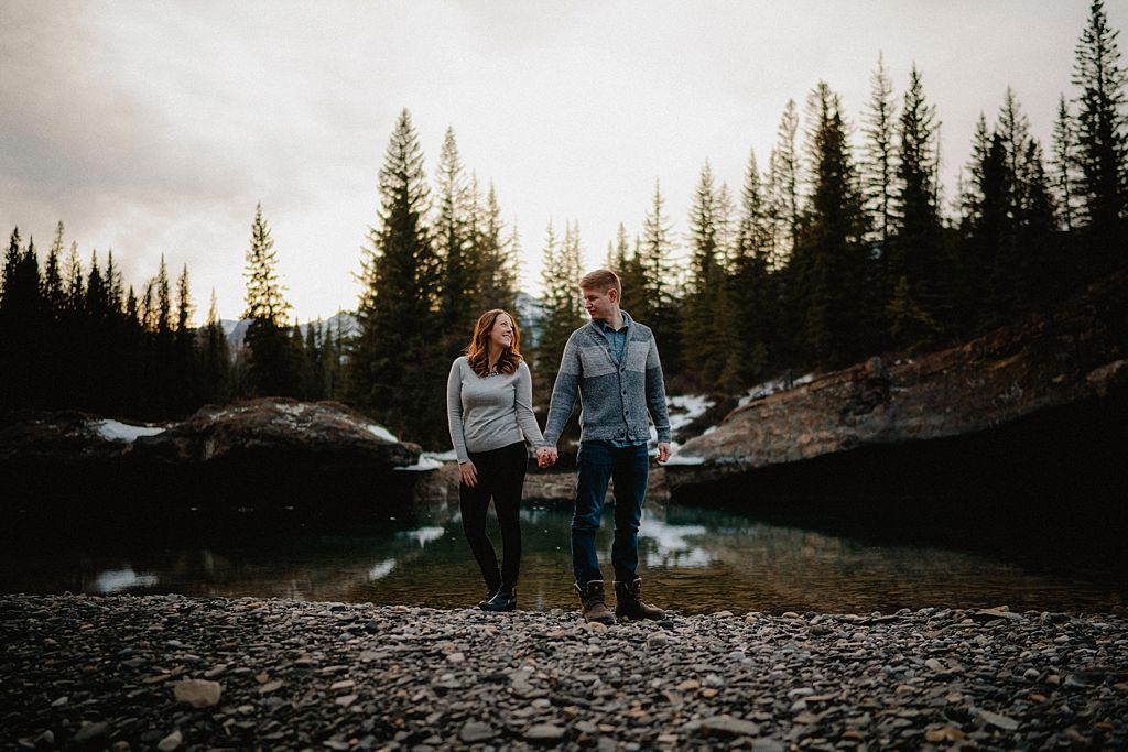 A couple walks along a rocky shoreline with evergreen trees silhouetted against evening light.