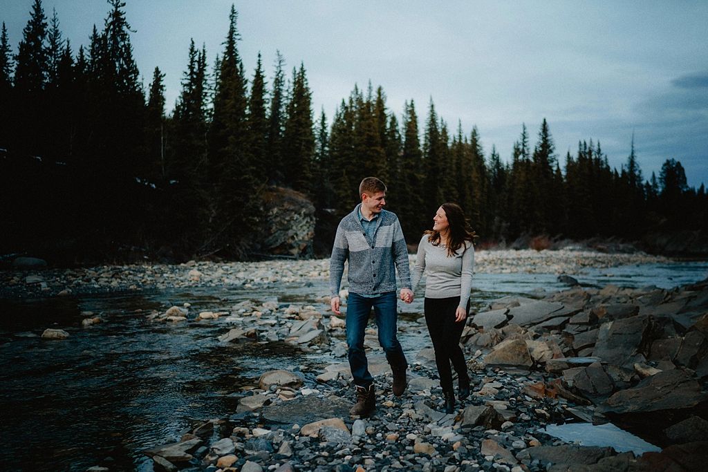 Couple walking together along a rocky shoreline with evergreen trees in the background.