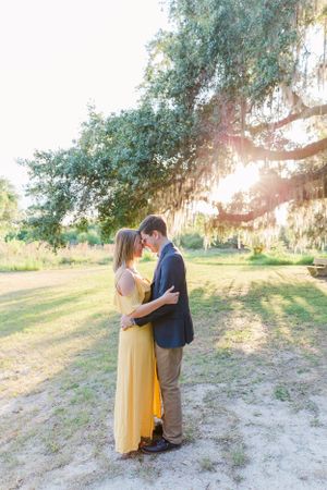Couple embracing in sunlit field with Spanish moss hanging from trees.