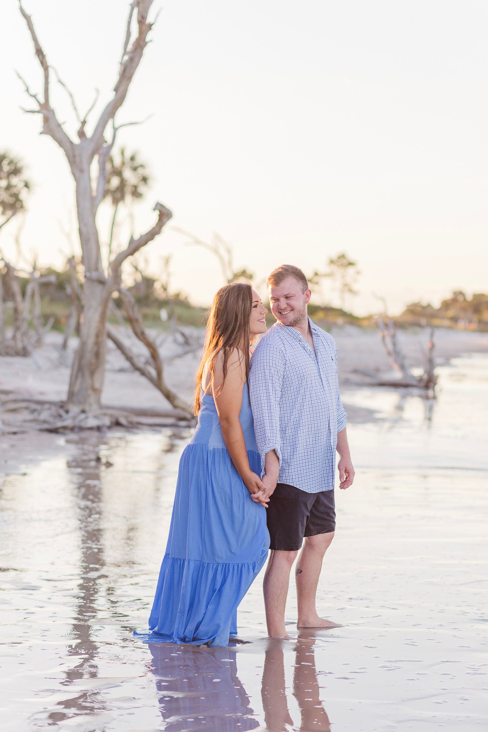 A couple walks along a beach at sunset with bare trees in the background creating a romantic silhouette.