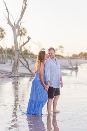 A couple walks along a beach at sunset with bare trees in the background creating a romantic silhouette.