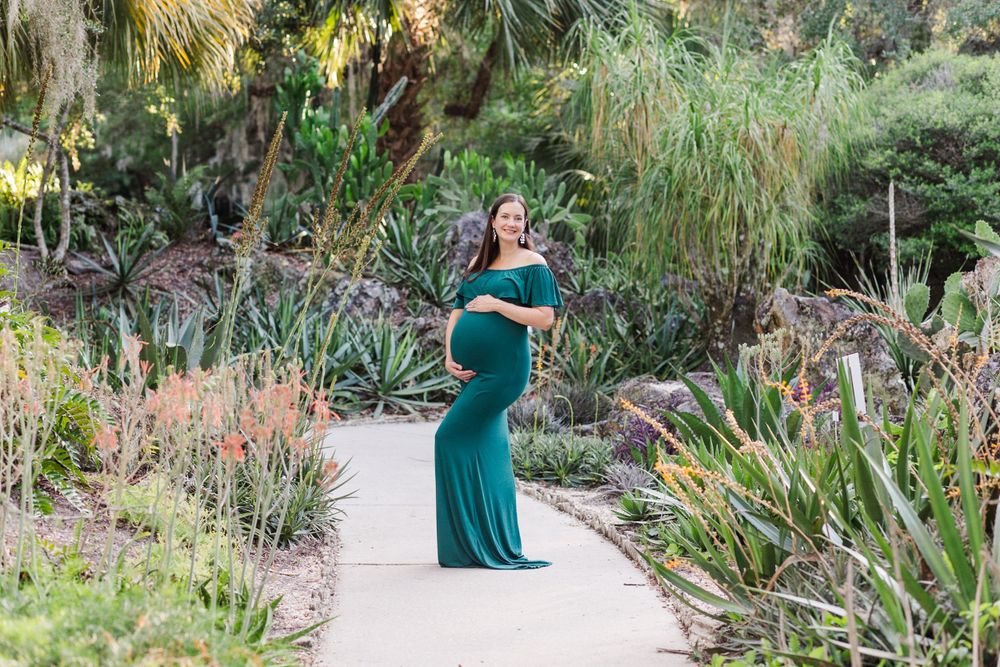 Maternity photo of woman in emerald green dress posing in tropical garden setting.