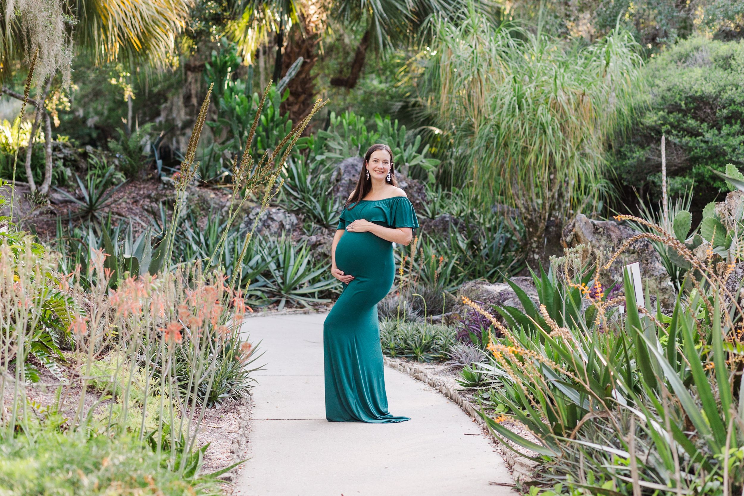 Maternity photo of woman in emerald green dress posing in tropical garden setting.