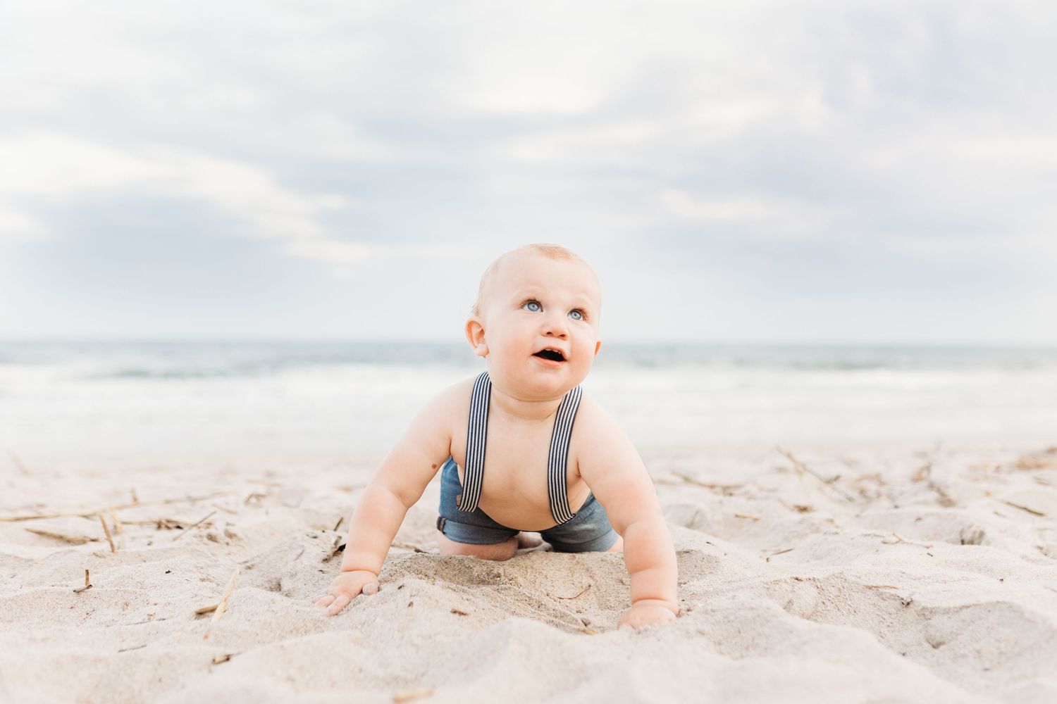 Small baby in gray overalls crawls happily on sandy beach with cloudy sky background.
