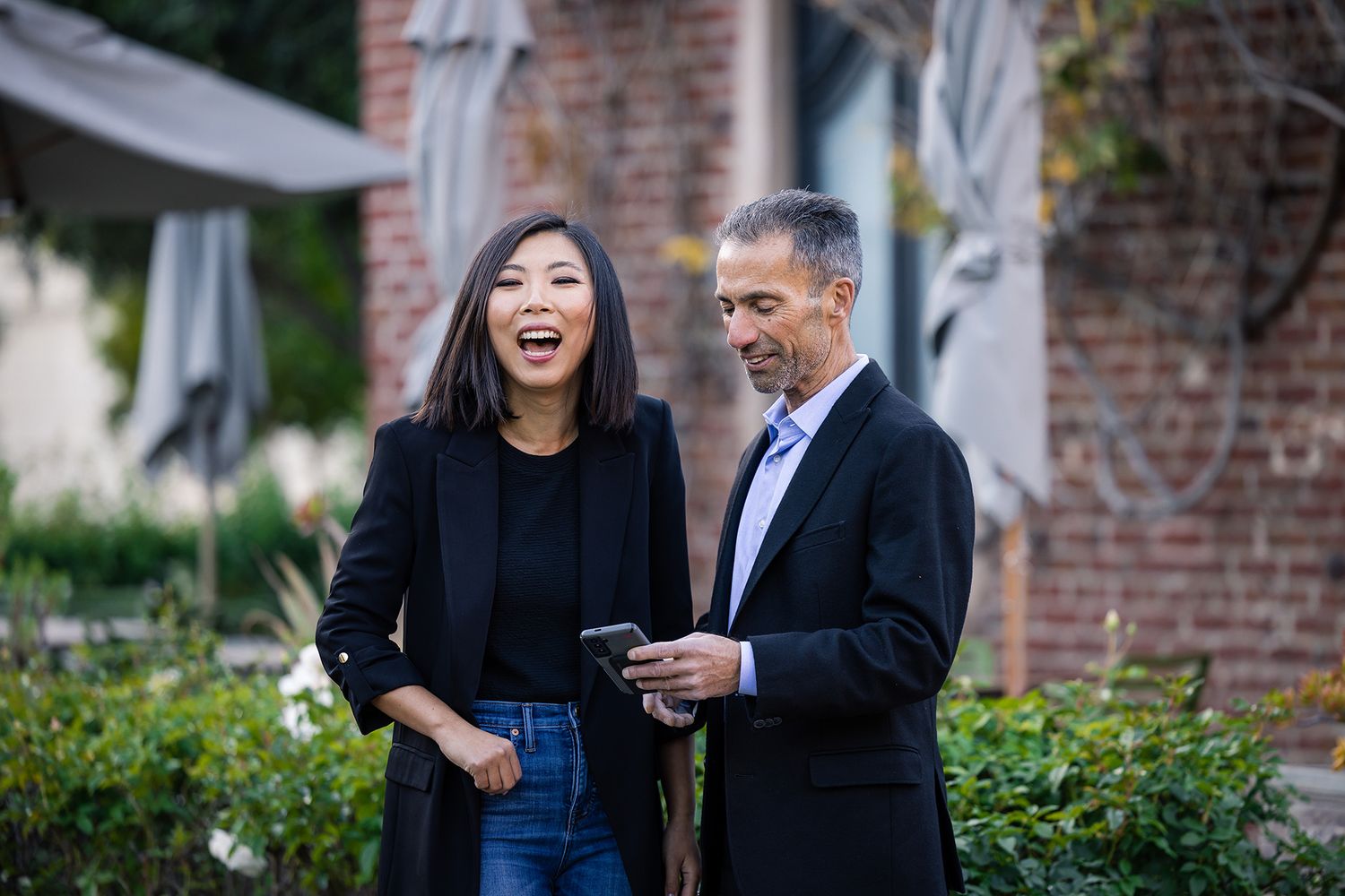Woman wearing black top and black jacket laughing and staring next to a man looking at his phone