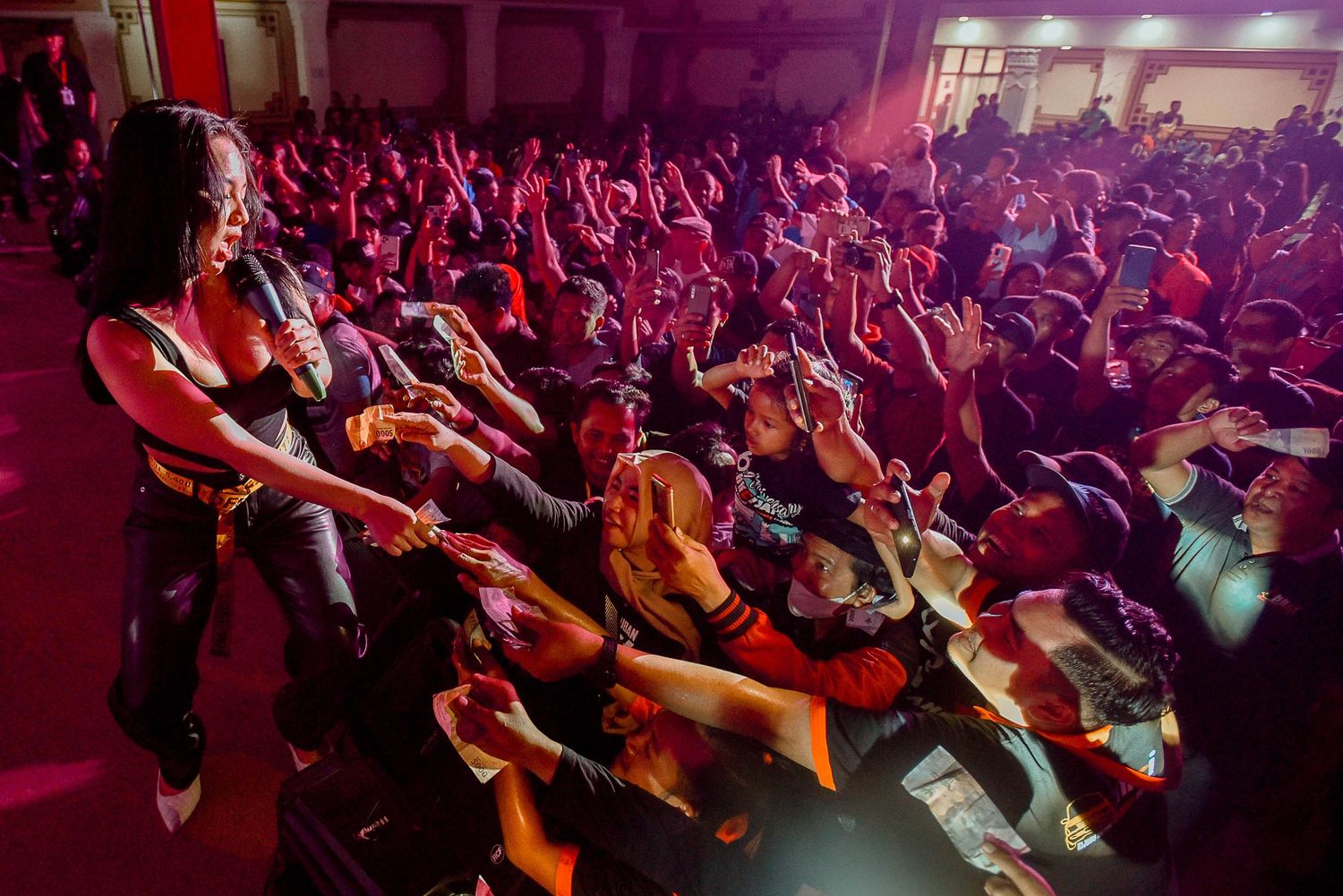 Concert performer engaging with a large crowd bathed in red stage lighting.