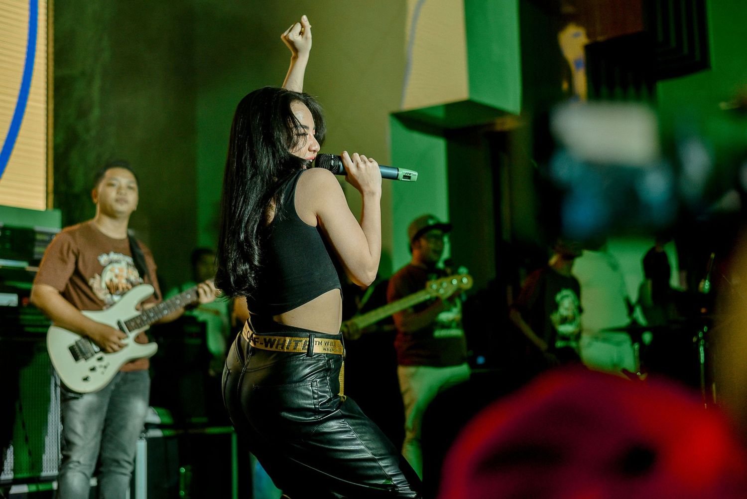 A rock band performs on stage with a singer in black outfit and guitarist under green lighting.