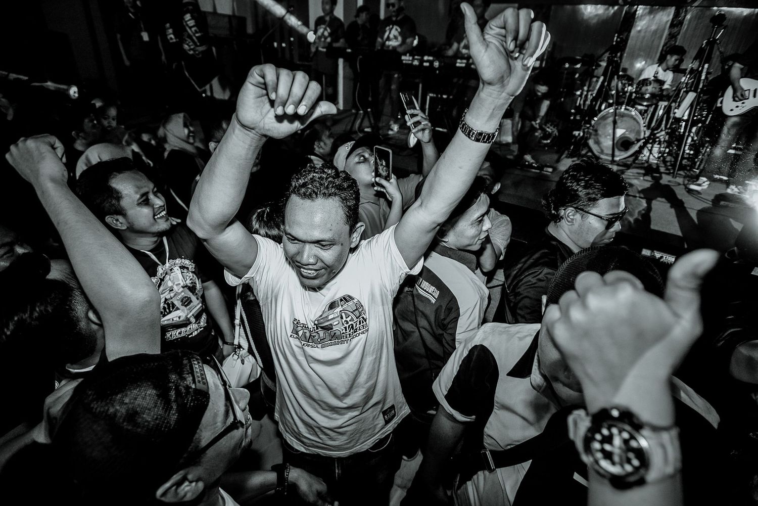 Black and white photo of enthusiastic concert-goers with raised arms in a packed venue.