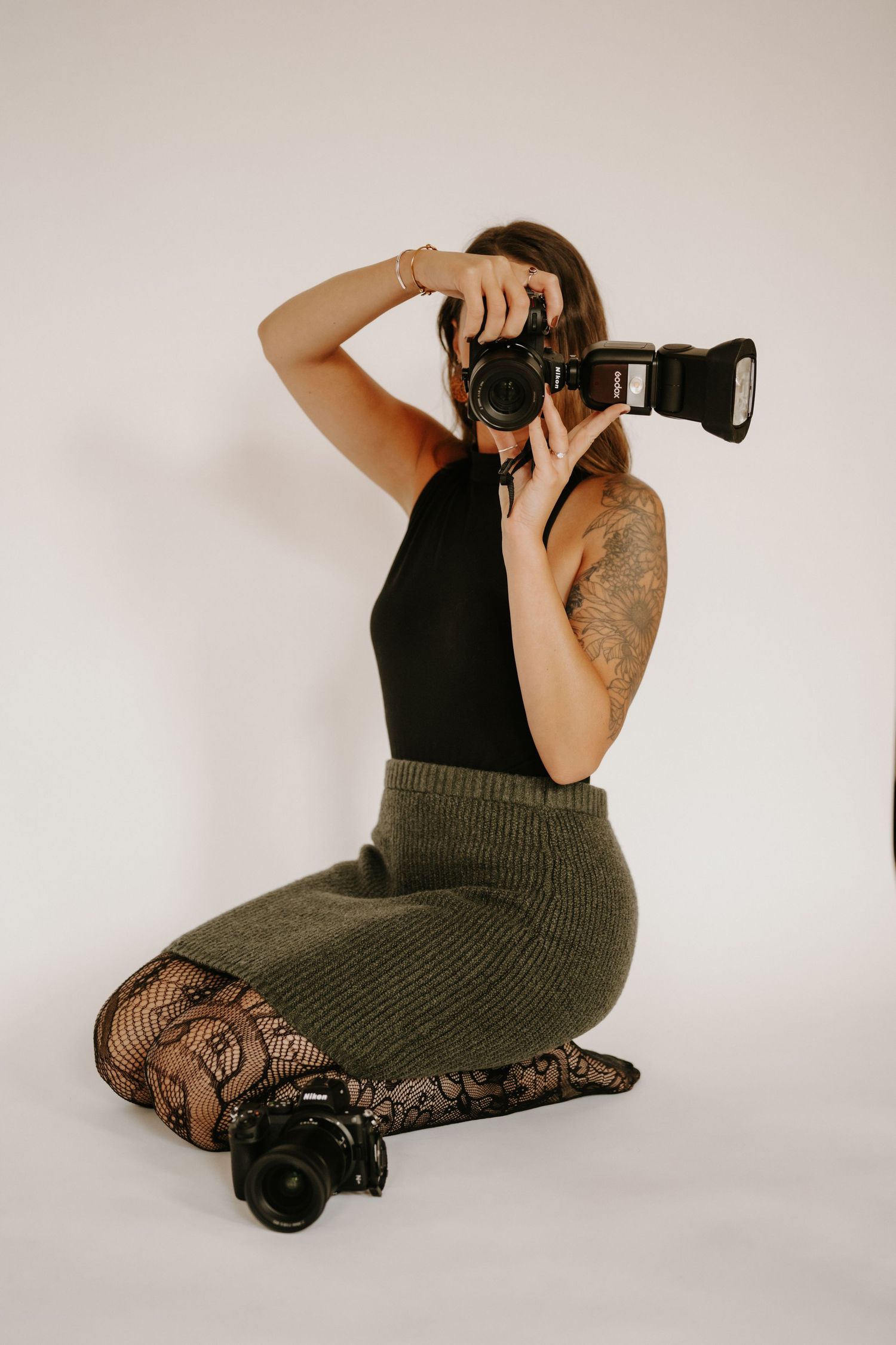 A photographer in dark clothing kneels with professional camera equipment against a light background.