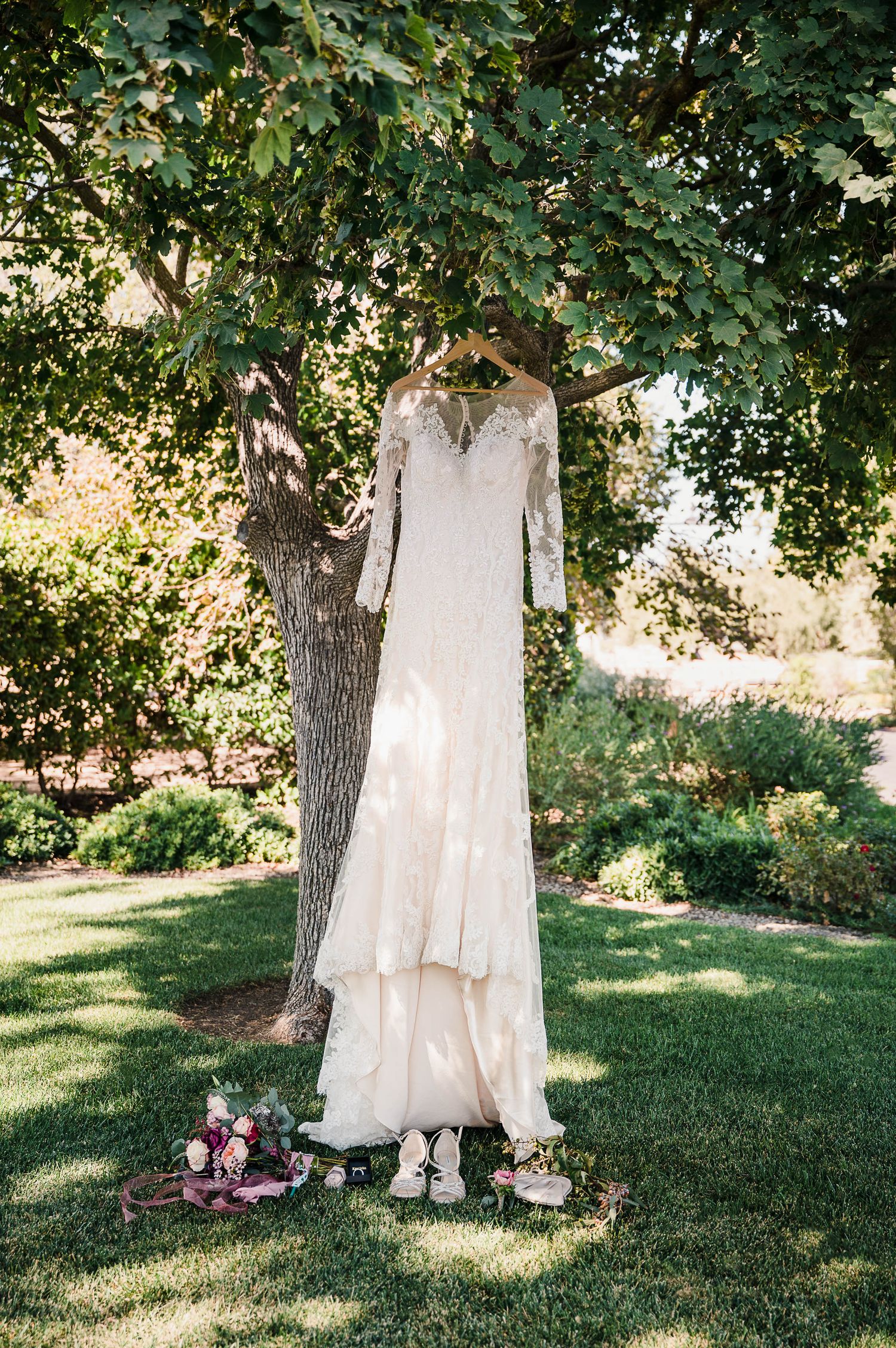 White lace wedding dress hanging from tree with bouquet and shoes beneath on grass.