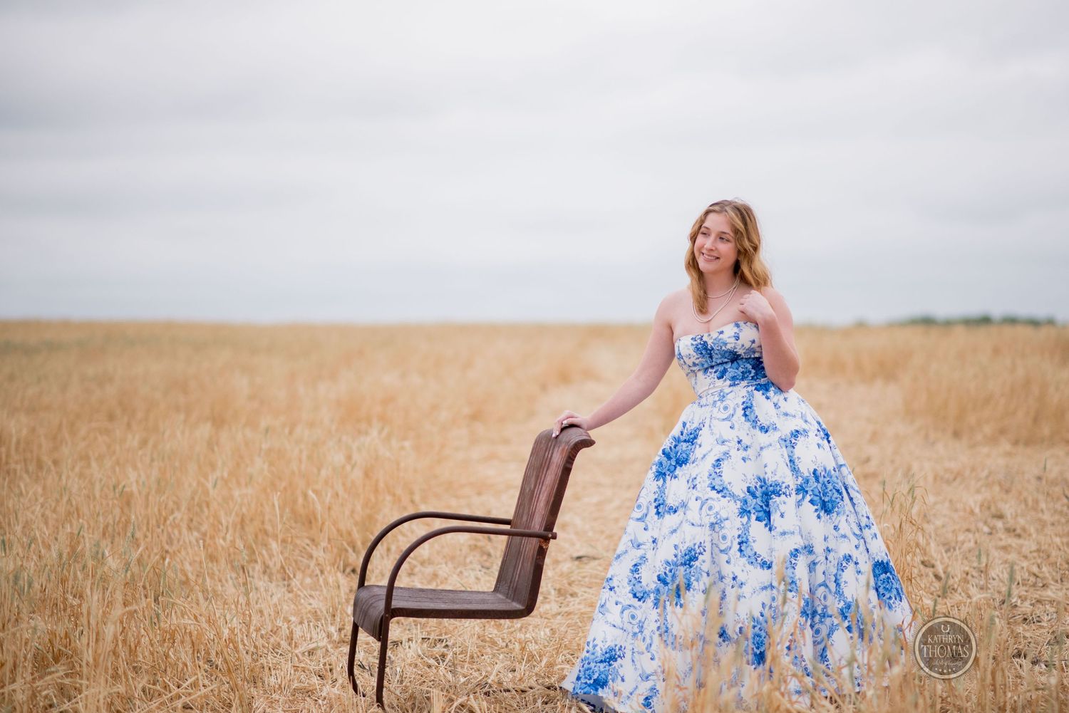 Elegant pose beside rustic chair in blue and white floral ballgown amid golden wheat field.