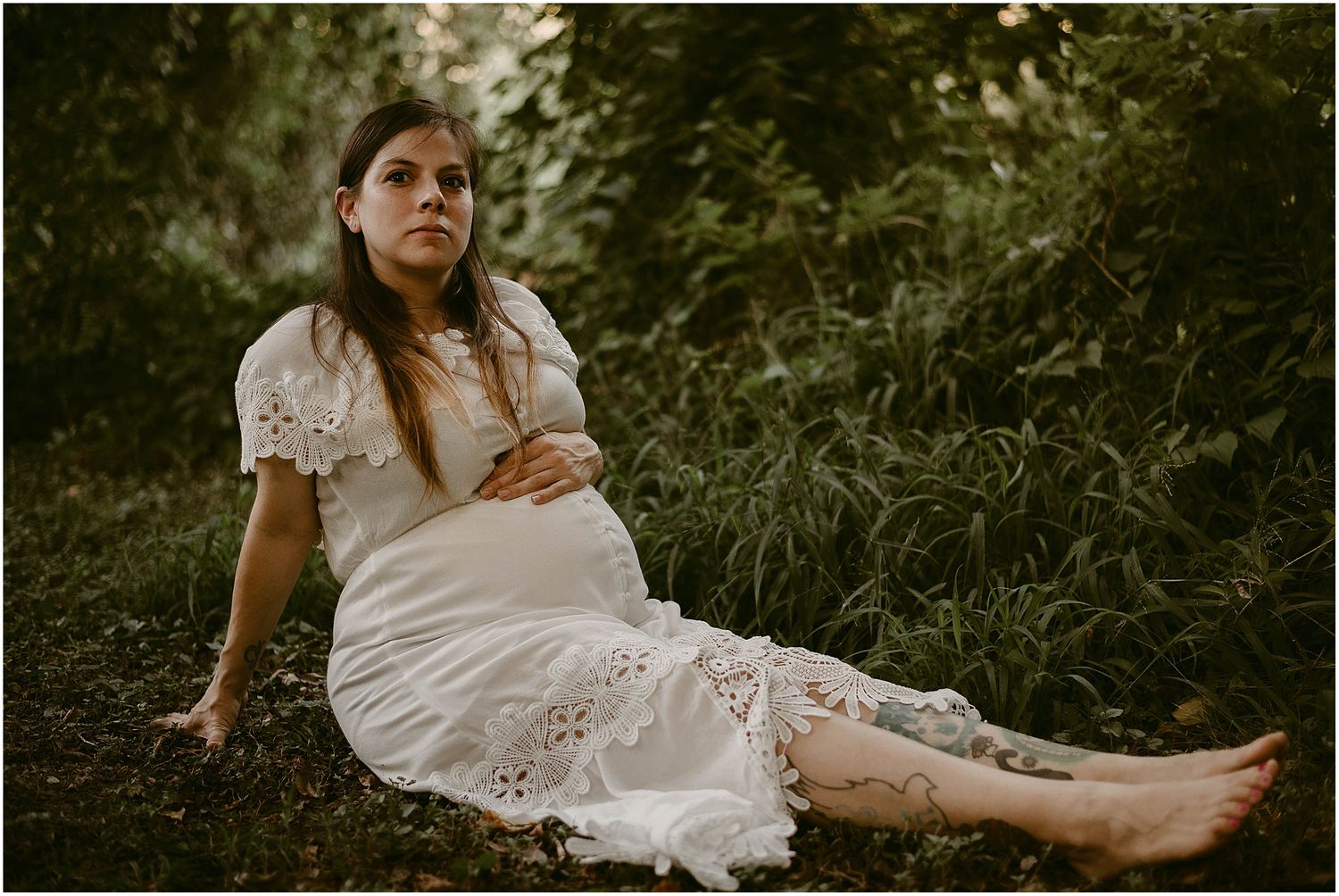 Expecting mother in vintage-style white lace dress rests gracefully on mossy ground during sunset.