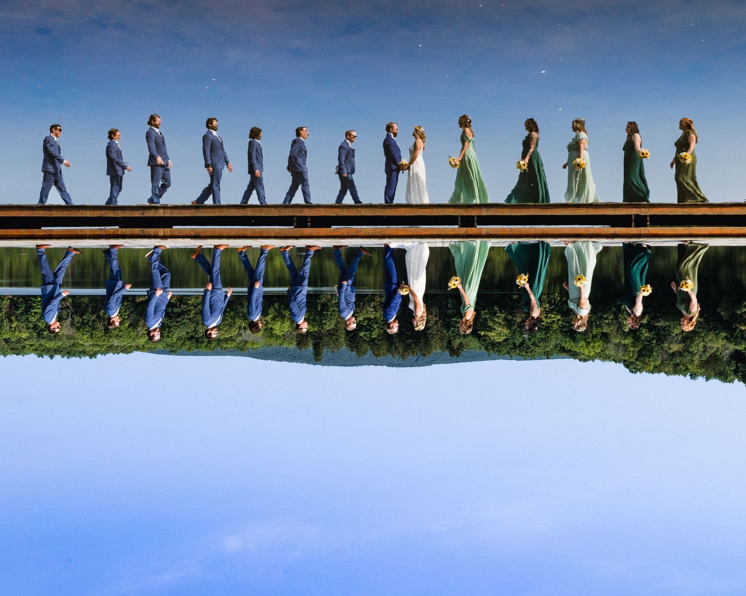 Wedding party standing in a line on bridge with perfect reflection in water below.