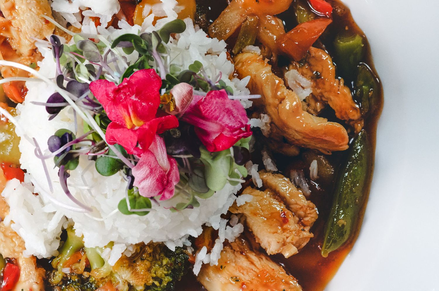 Asian stir-fry dish with chicken, vegetables, steamed rice, and fresh pink edible flower garnish.