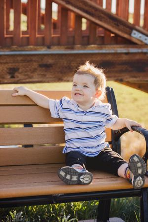 Small child in striped shirt sits on wooden bench during sunny afternoon.