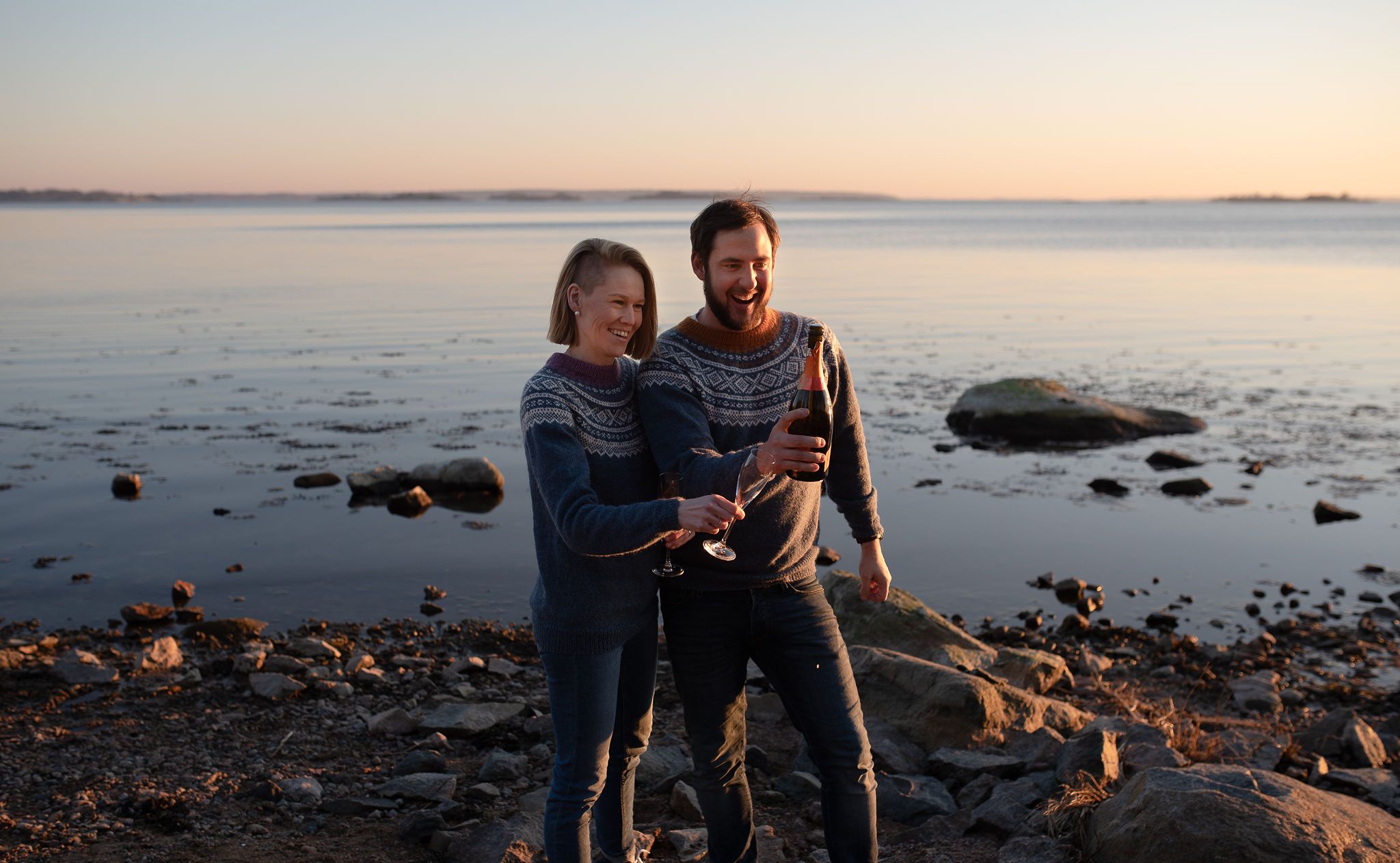 Couple in matching sweaters embrace by rocky beach at sunset.