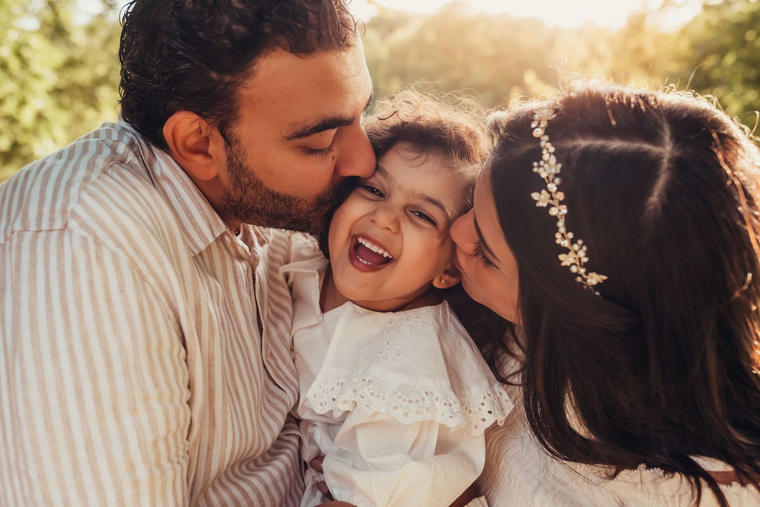 parents kissing their daughter on the cheek, laughing, sunset, white dress