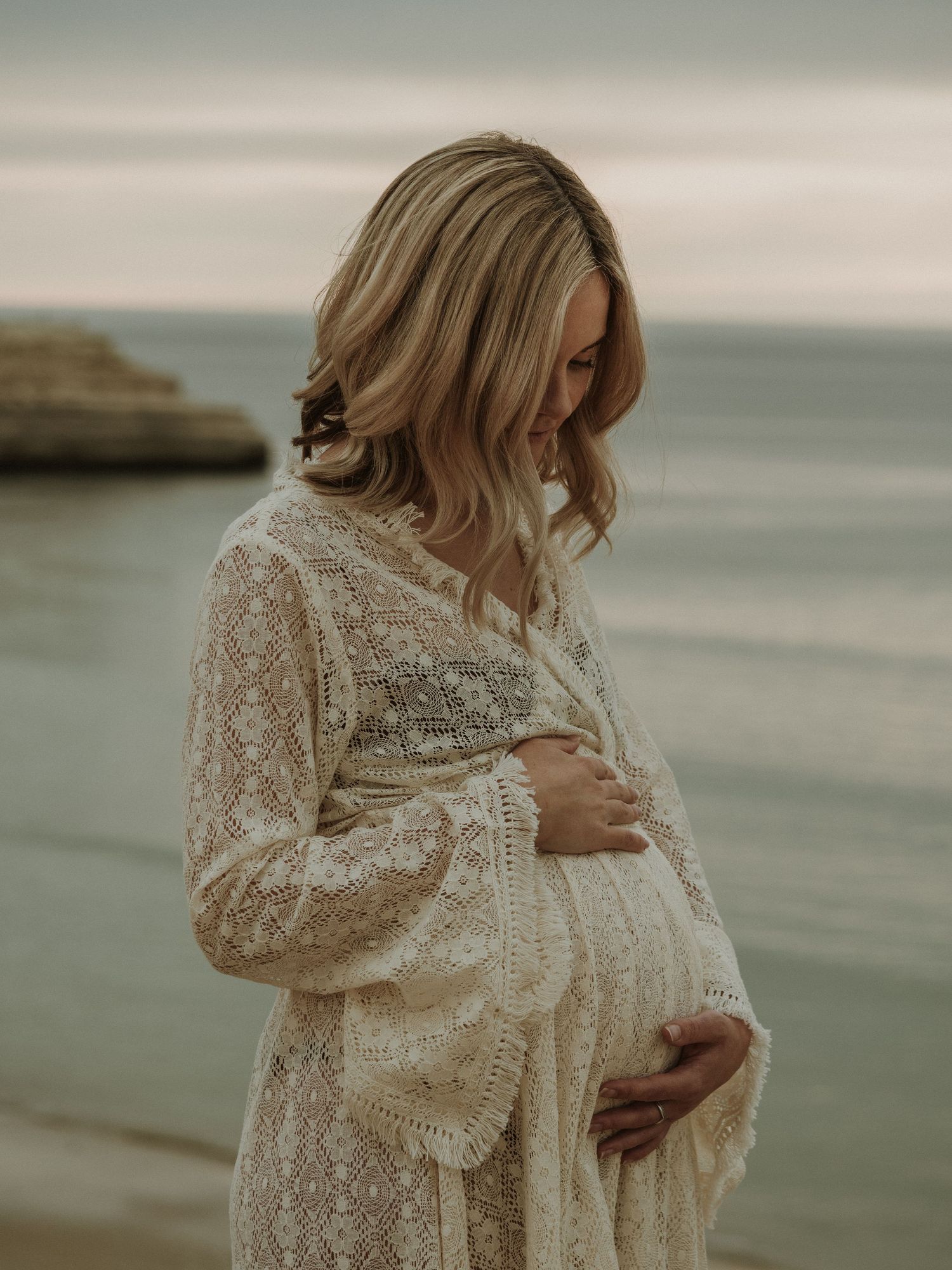 Maternity portrait featuring a white lace dress against coastal cliffs at sunset.