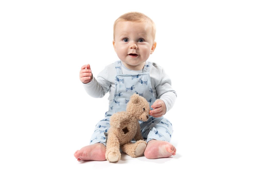 A boy toddler sits wearing a grey top and blue dungarees and holding a brown teddy, against a white backdrop.