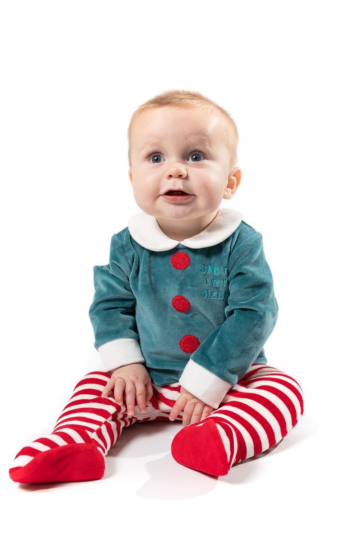 A boy toddler sits against a white backdrop wearing a Christmas outfit of a green top and red and white striped bottoms.