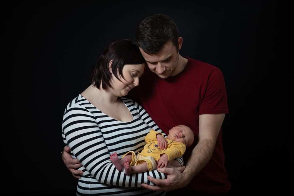 Young parents stand, cradling their newborn son between them as they look towards him.