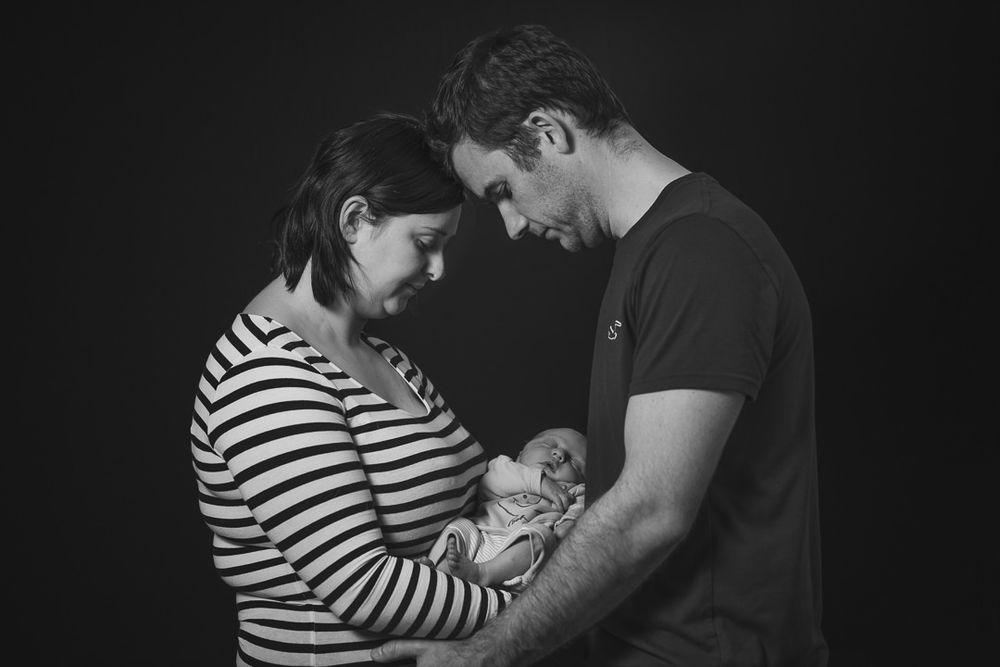 Young parents stand face to face, resting their foreheads together while cradling their newborn son in the middle.
