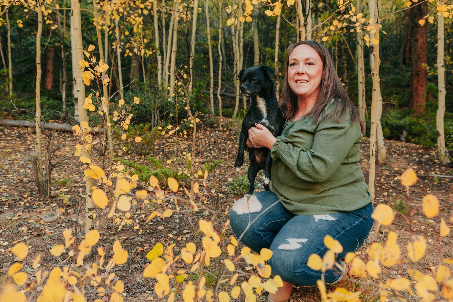 Someone in a green jacket poses with their black dog amid yellow fall leaves.